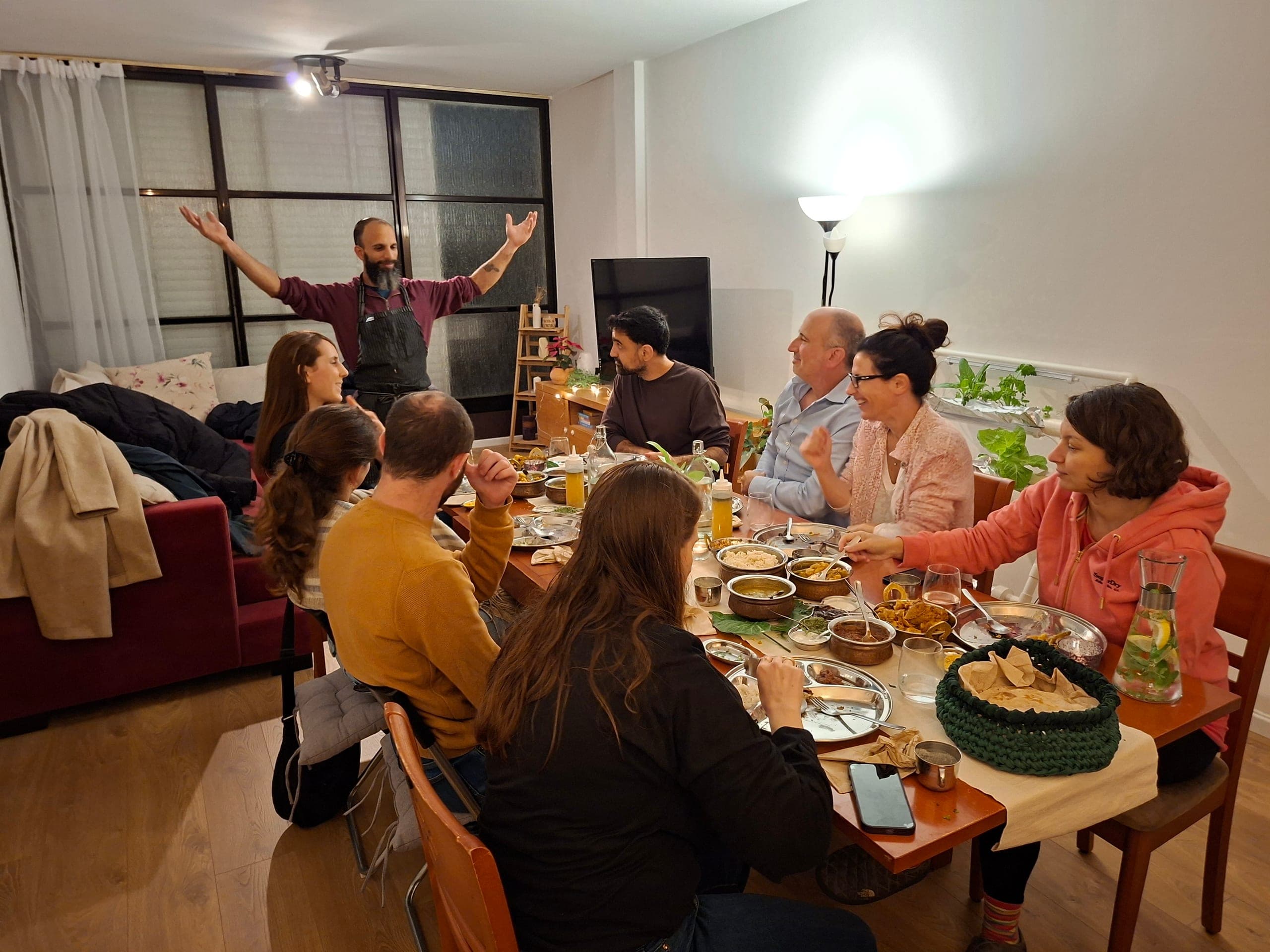 קבוצת אנשים יושבת סביב שולחן ארוך ונהנית מארוחה הודית עשירה, עם מנות רבות בכלי הגשה מסורתיים. אחד הגברים עומד ומדבר בהתלהבות עם ידיים פרושות.